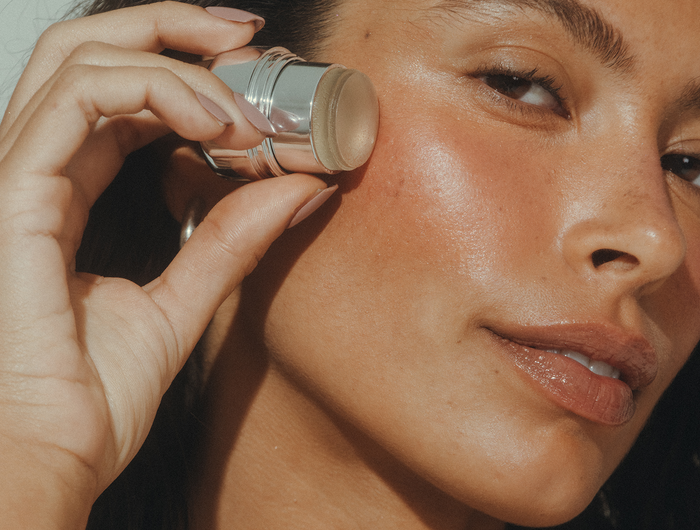 A close-up lifestyle shot of a woman applying a beige makeup stick, likely a contour or bronzer, onto her cheek. The product container is silver and clear, showing the beige stick tip.